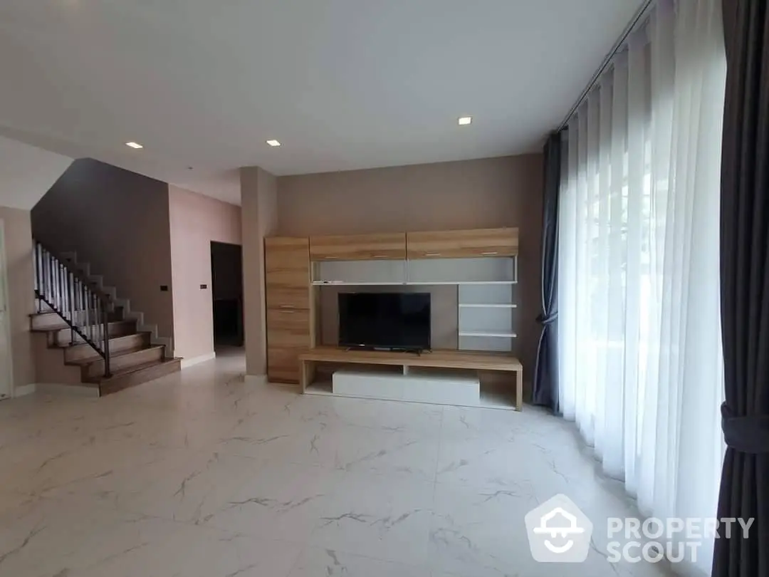 Spacious living room with elegant marble flooring, modern built-in entertainment unit, and large windows draped with luxurious curtains, complemented by a stylish staircase.