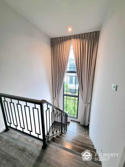 Elegant staircase with modern railing and large window with curtains in a bright home interior.