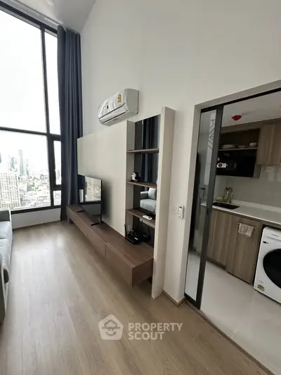 Modern living room with high ceilings and city view, adjacent to a sleek kitchen with appliances.