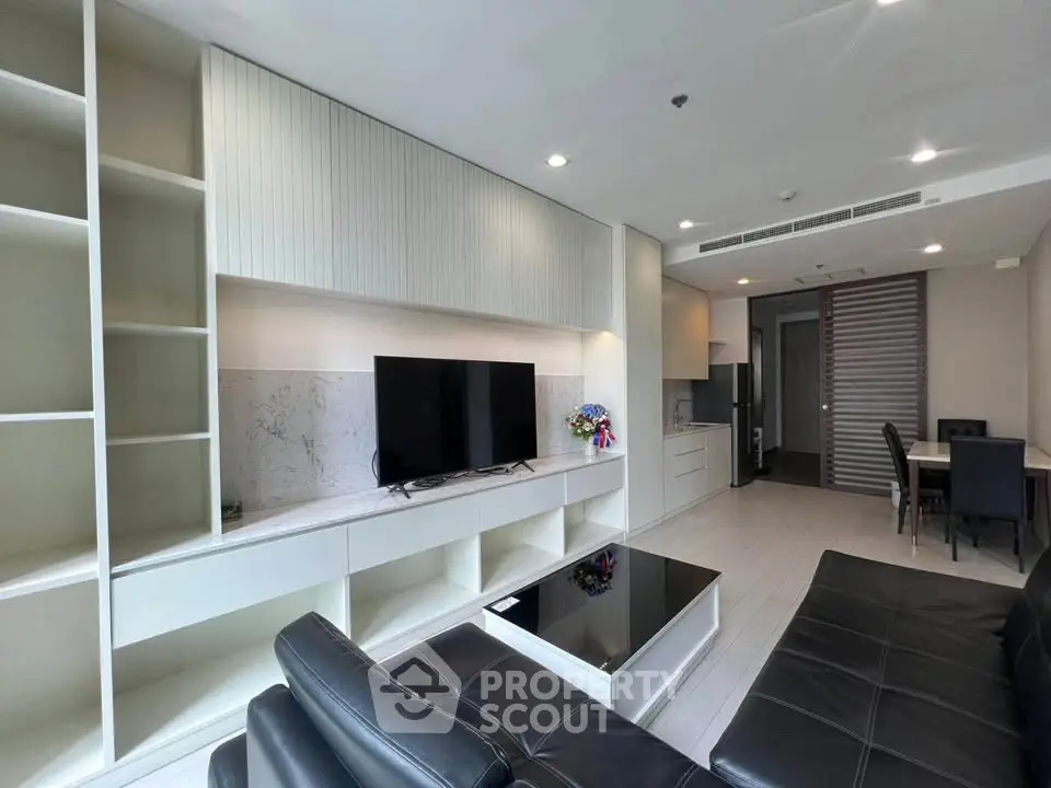 Modern living room with sleek black leather sofas and built-in shelving, open to a stylish kitchen area.