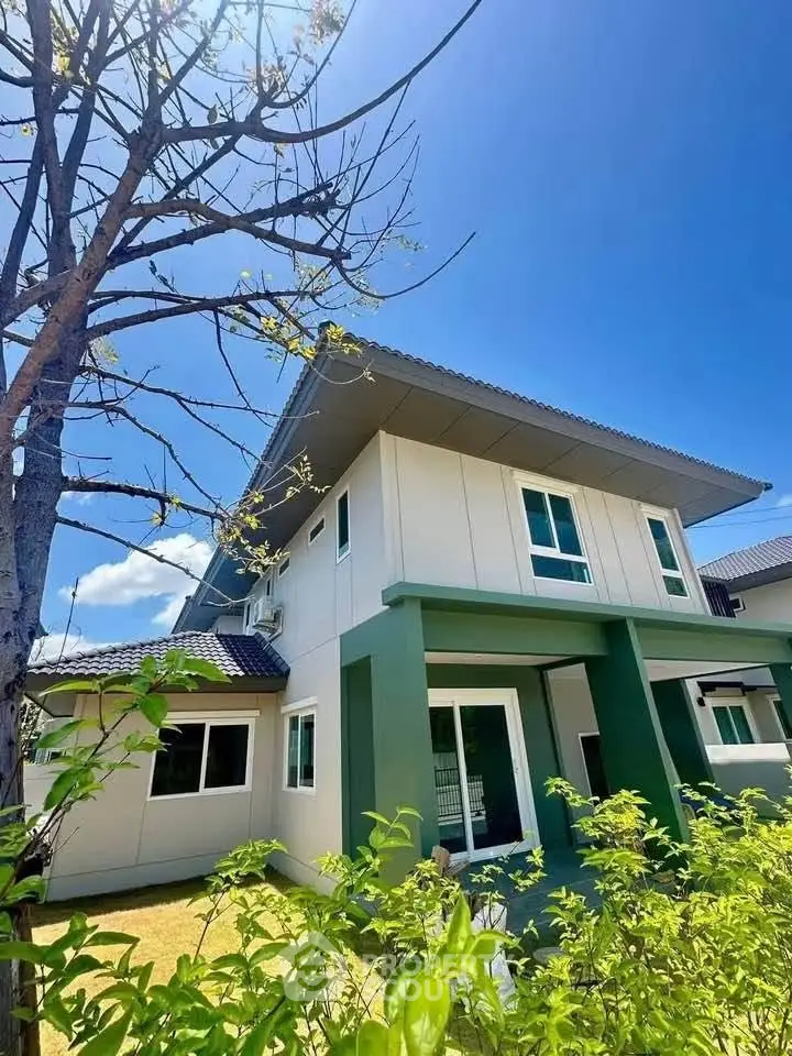 Modern two-story house with green accents and lush garden under clear blue sky.