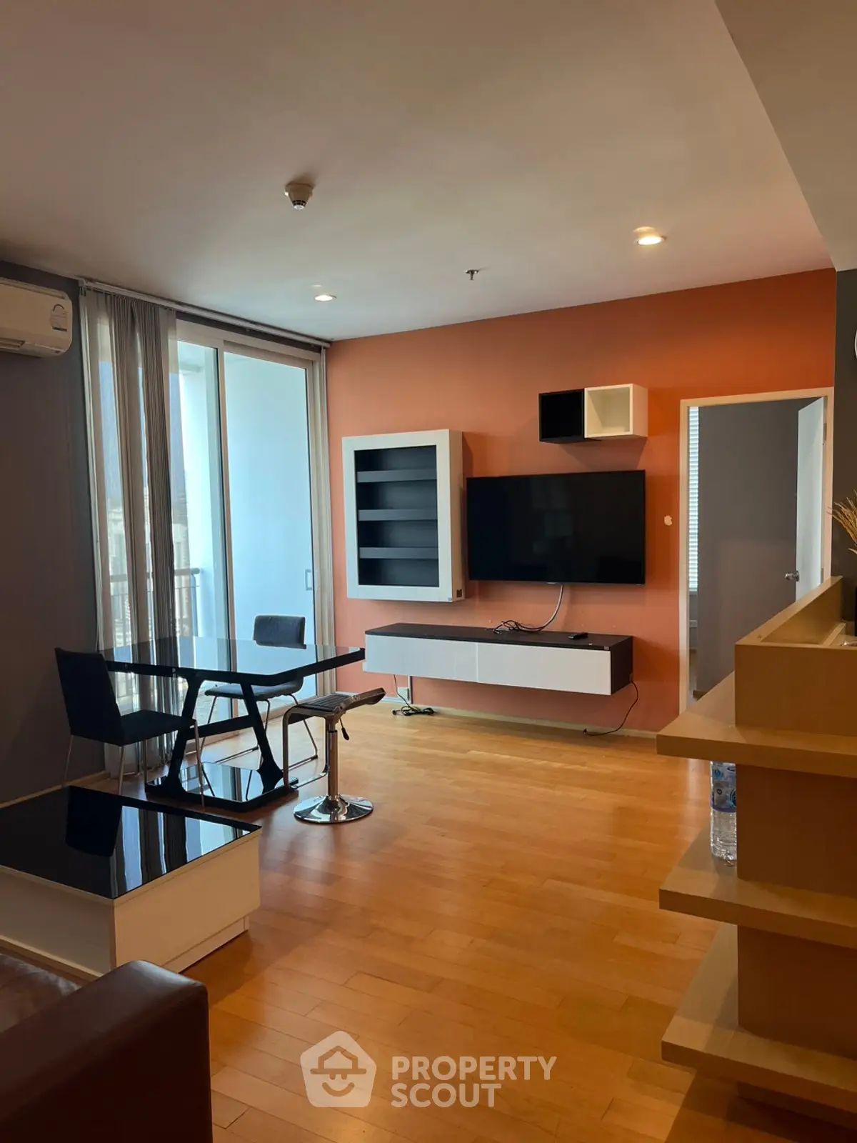 Modern living room with sleek furniture and large TV, featuring wooden flooring and balcony access.
