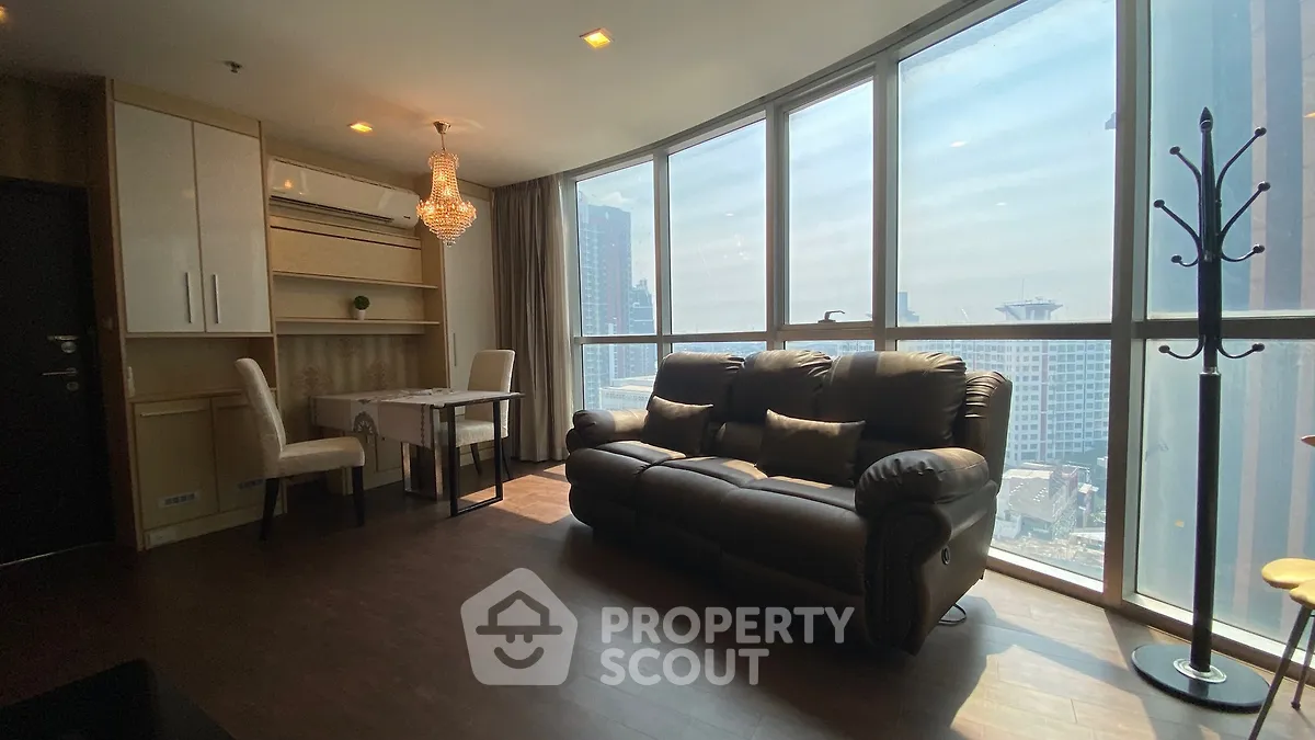 Spacious living room with large windows and city view, featuring elegant chandelier and cozy seating.