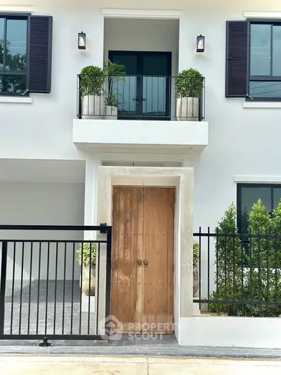 Modern two-story house with elegant entrance and balcony, featuring lush greenery and sleek design.