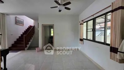 Spacious living room with ceiling fan and tiled flooring, featuring large windows and staircase.