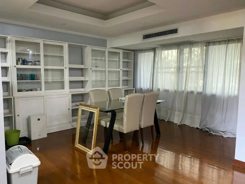Spacious dining area with glass table and elegant chairs, featuring large windows and wooden flooring.