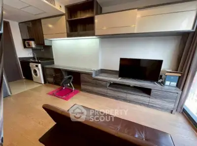 Modern living room with integrated kitchen and workspace, featuring sleek cabinetry and a washing machine.
