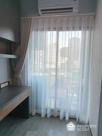 Sunlit modern bedroom with floor-to-ceiling sheer curtains, offering a glimpse of the cityscape, complemented by sleek furniture and an air conditioning unit.