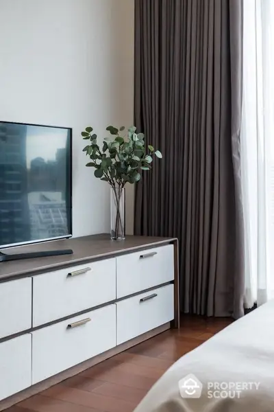 Modern bedroom with sleek dresser and large TV, complemented by elegant curtains and stylish decor.