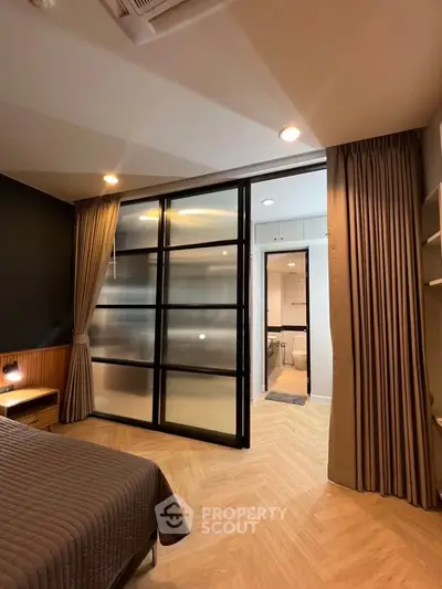 Modern bedroom with sliding glass doors leading to a stylish bathroom, featuring elegant lighting and wood flooring.