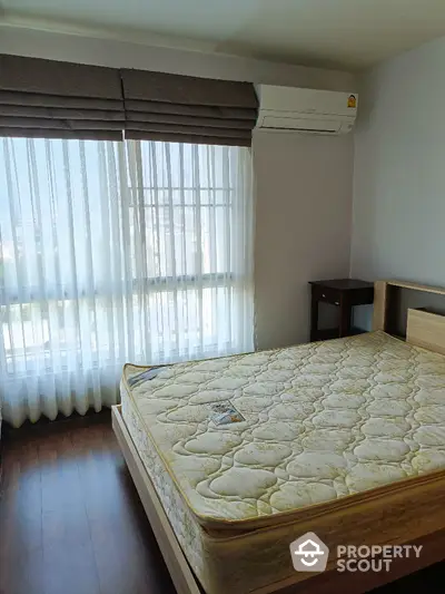 Bright bedroom with large window and air conditioning unit, featuring a comfortable bed and wooden flooring.