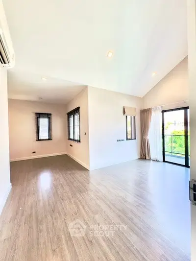 Spacious modern living room with large windows and balcony access, featuring sleek wooden flooring and ample natural light.