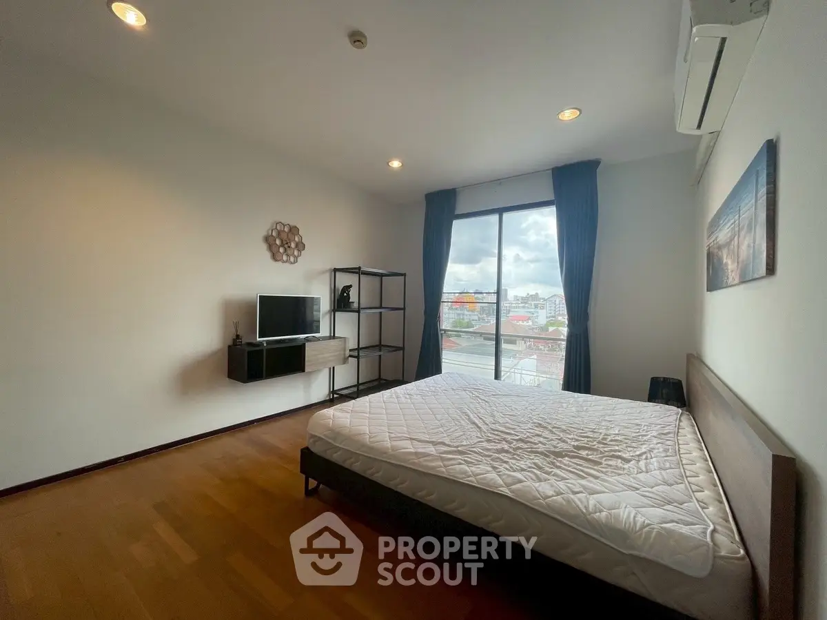 Spacious bedroom with large window and city view, featuring modern furniture and wooden flooring.