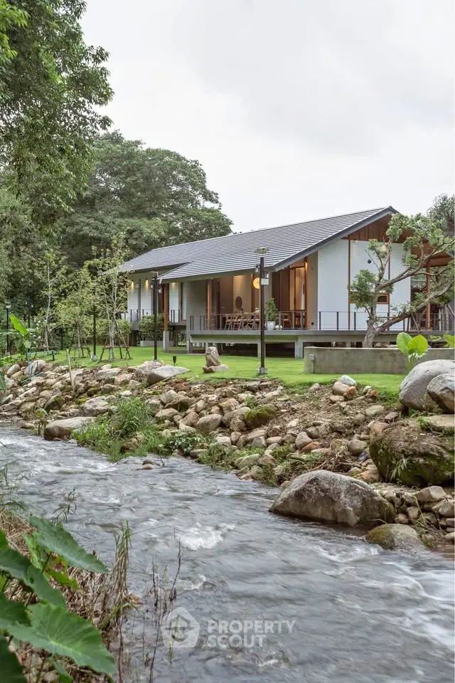 Charming modern house with spacious porch by a serene creek, surrounded by lush greenery.