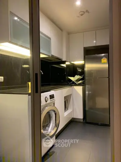 Modern kitchen with sleek black countertops and built-in appliances, featuring a washing machine and stainless steel fridge.