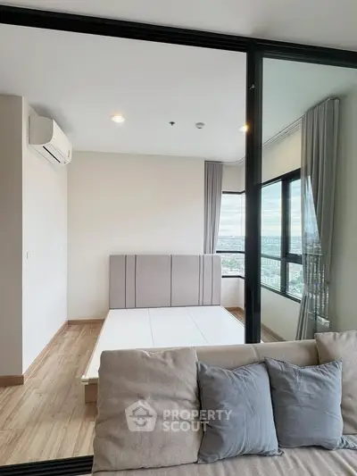 Modern bedroom with large windows and city view, featuring a minimalist bed frame and cozy seating area.