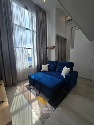 Modern living room with high ceiling and blue sofa, featuring large windows and natural light.