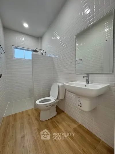Modern bathroom with sleek white tiles and wooden flooring, featuring a shower, toilet, and sink.