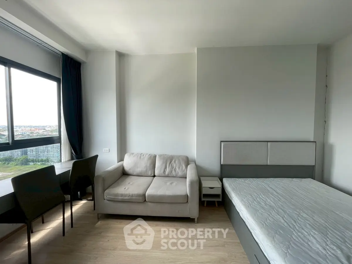 Modern studio apartment with cozy sofa and bed, featuring large window with city view.
