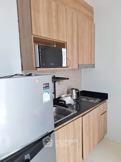 Modern compact kitchen with wooden cabinets, microwave, and stainless steel fridge