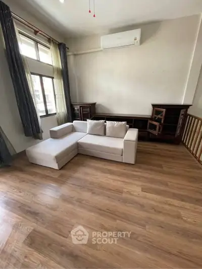 Spacious living room with modern sofa and wooden flooring, featuring large windows and air conditioning.