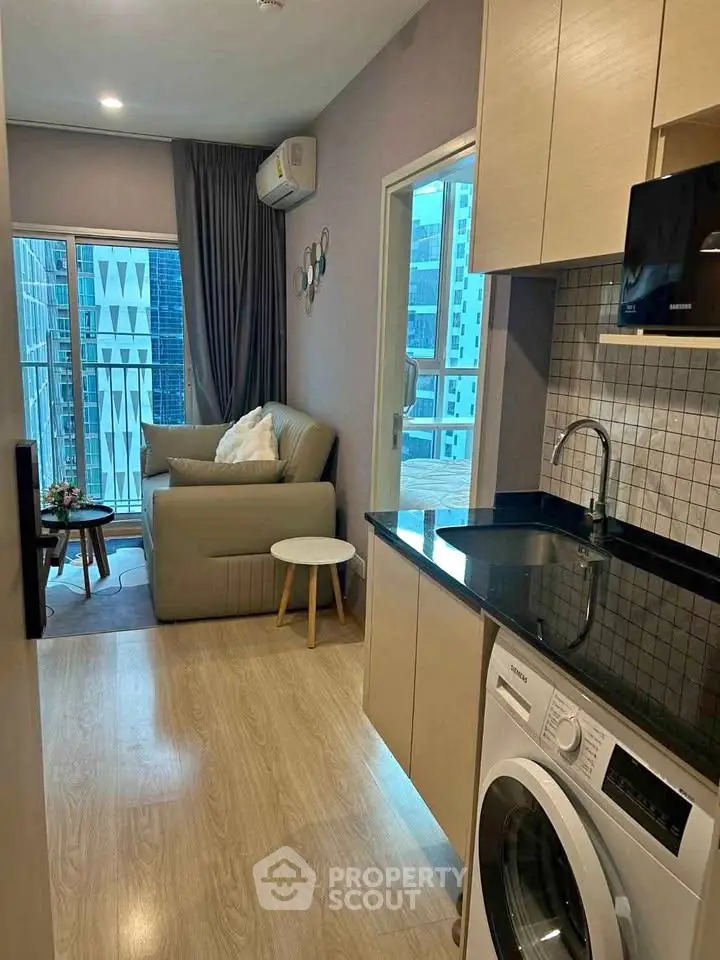 Modern apartment interior with compact kitchen and cozy living area, featuring a washing machine and city view.