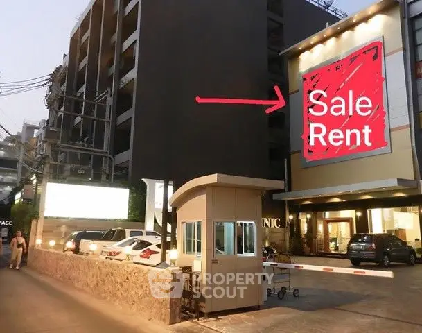 Modern apartment building exterior with sale and rent sign, featuring parking area and security booth.