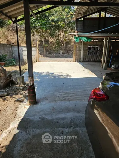 Spacious outdoor parking area with a protective metal roof, surrounded by natural greenery, offering ample space for multiple vehicles and outdoor activities.