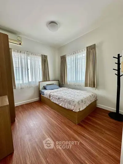 Cozy bedroom with wooden flooring and natural light from two windows, featuring a single bed and minimalist decor.