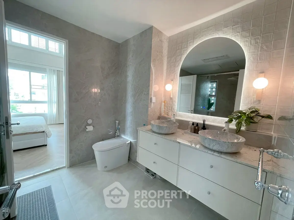 Luxurious bathroom with dual sinks and elegant marble finishes, leading to a bright bedroom with large windows.