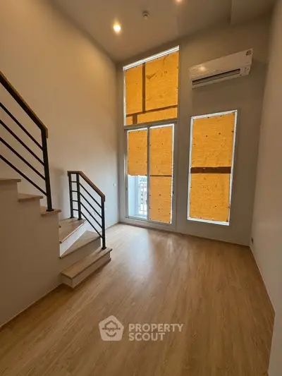 Modern duplex living room with high ceilings and large windows, featuring a sleek staircase and wooden flooring.