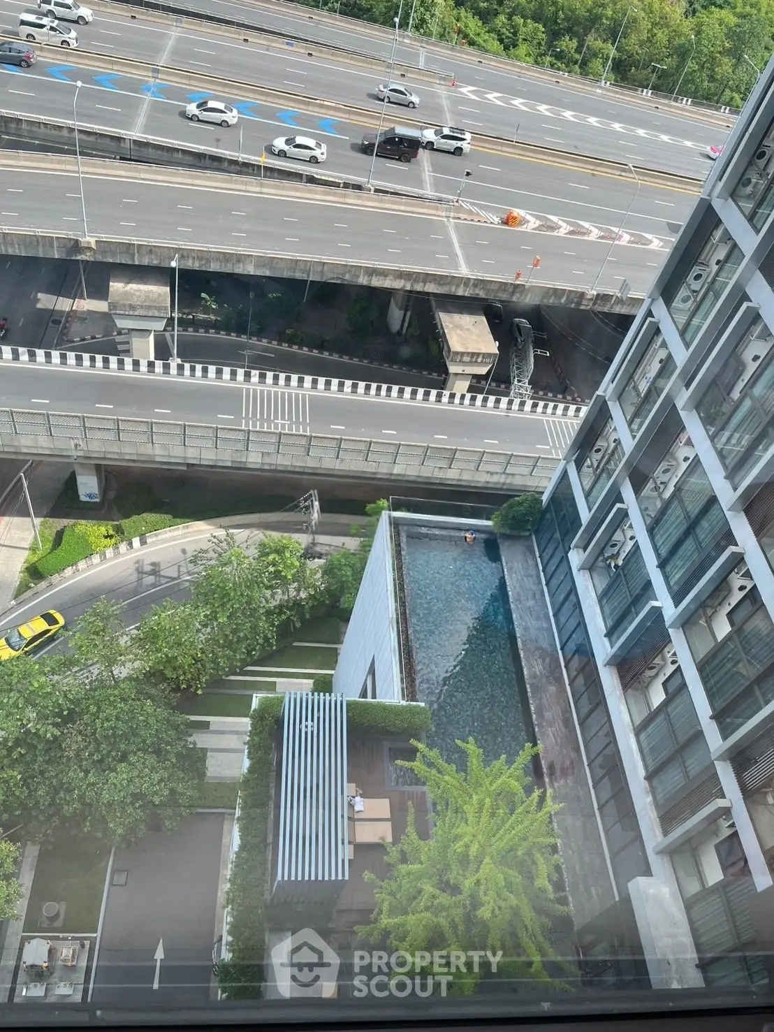 Stunning aerial view of modern building with rooftop pool and lush greenery, overlooking a busy highway.