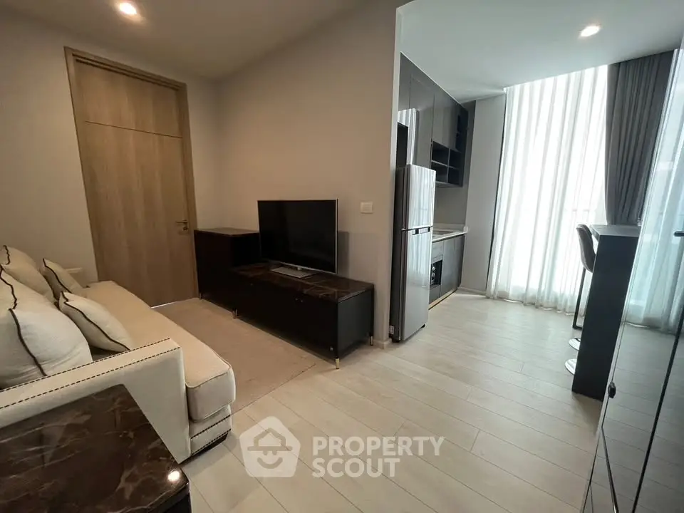 Modern living room with sleek kitchen, featuring elegant furniture and natural light from large windows.