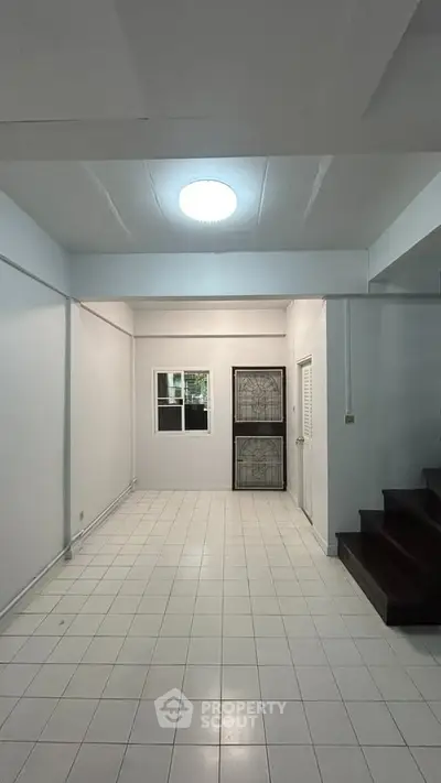 Spacious white-tiled room with staircase and window, ideal for versatile living space.