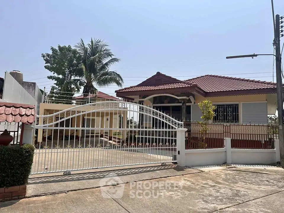 Charming single-story house with red roof and gated entrance, surrounded by lush greenery and palm trees.