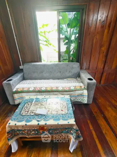 Cozy living room with wooden walls and floor, featuring a comfortable sofa and a window view of lush greenery.