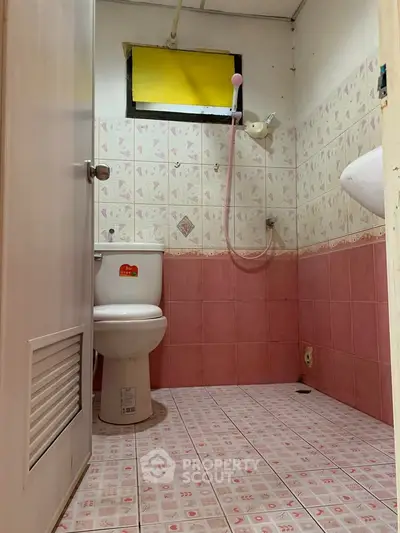Compact bathroom with pink tiles and showerhead, featuring a small window and basic fixtures.