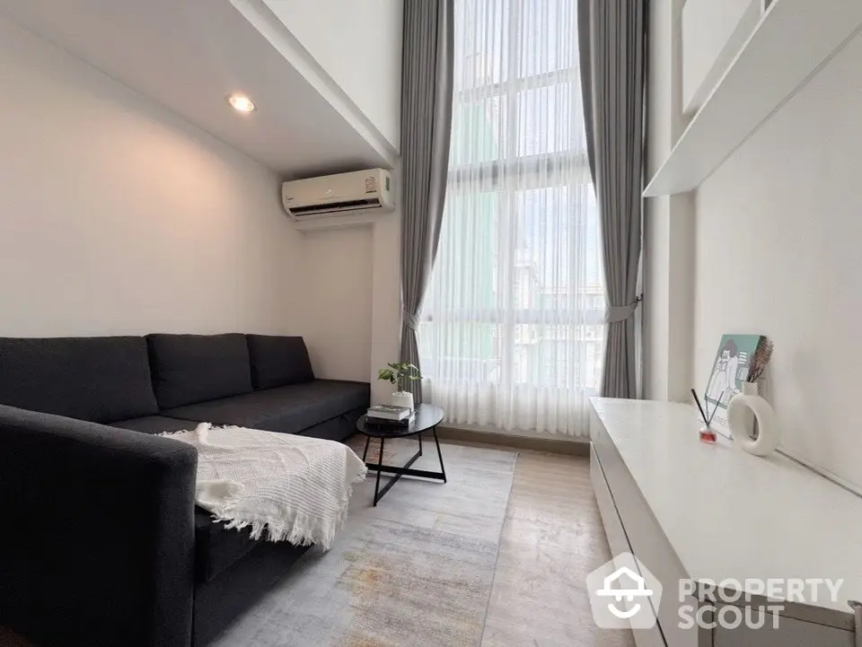 Modern living room with high ceiling and large window, featuring a sleek black sofa and minimalist decor.