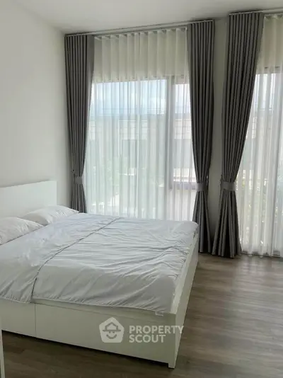 Bright and airy bedroom with large windows and elegant curtains, featuring a cozy bed and wooden flooring.