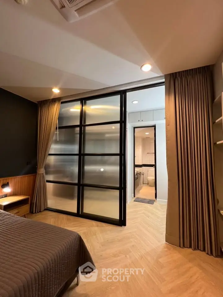 Modern bedroom with sliding glass doors leading to a stylish bathroom, featuring elegant lighting and wood flooring.