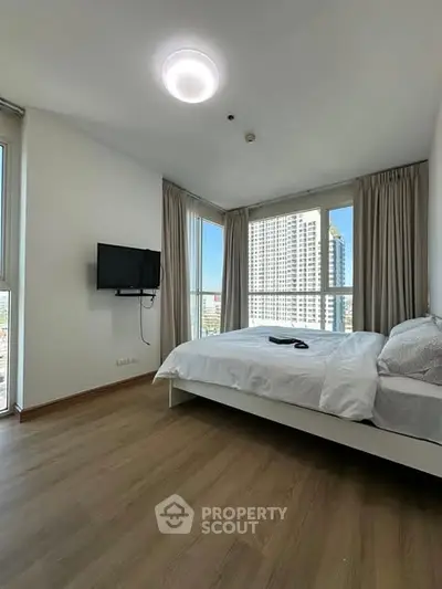 Modern bedroom with large windows and city view, featuring a wall-mounted TV and wooden flooring.