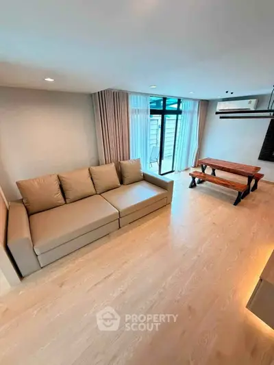 Spacious living room with modern sofa and dining area, featuring large windows and wooden flooring.