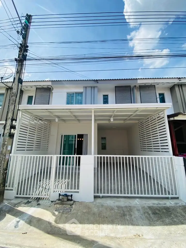 Modern townhouse exterior with white fence and covered parking area under clear blue sky.