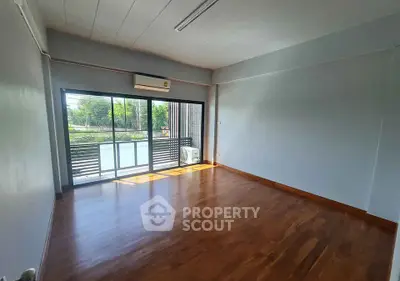 Spacious empty room with wooden flooring and large window overlooking greenery.