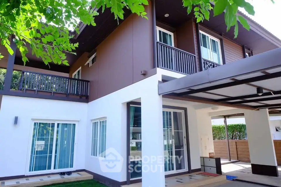 Modern two-story house with large windows and balcony, surrounded by lush greenery.