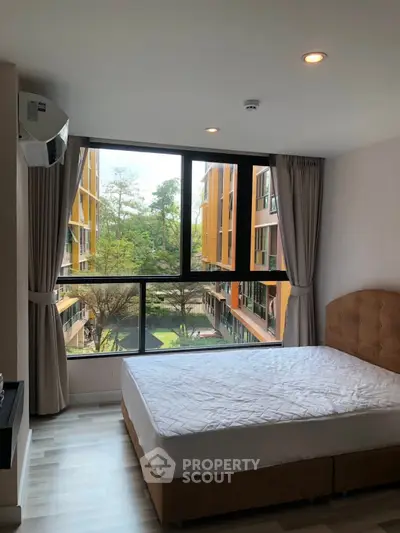 Bright bedroom with large window offering a stunning view of modern apartment complex and lush greenery.