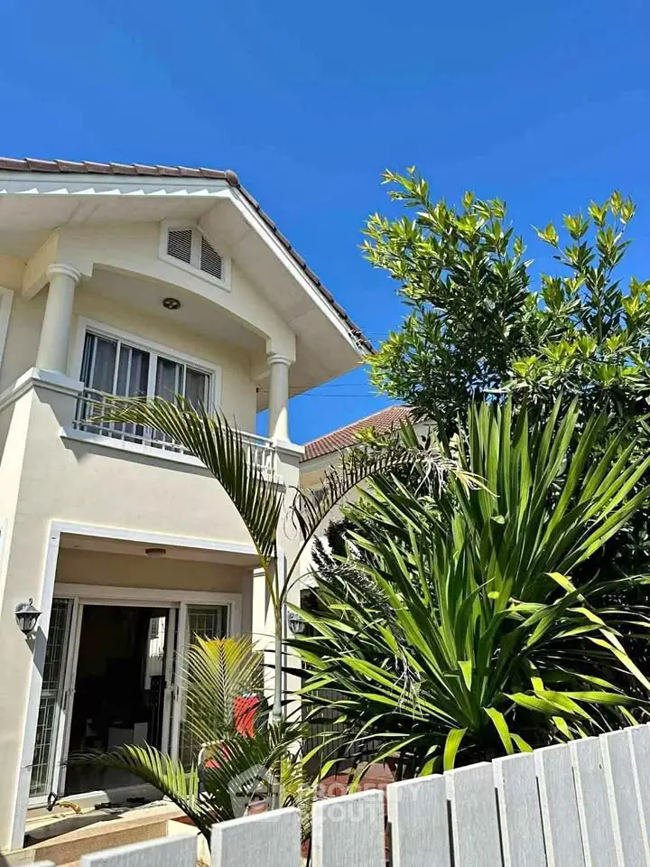 Charming two-story house with lush garden and elegant balcony under clear blue sky.