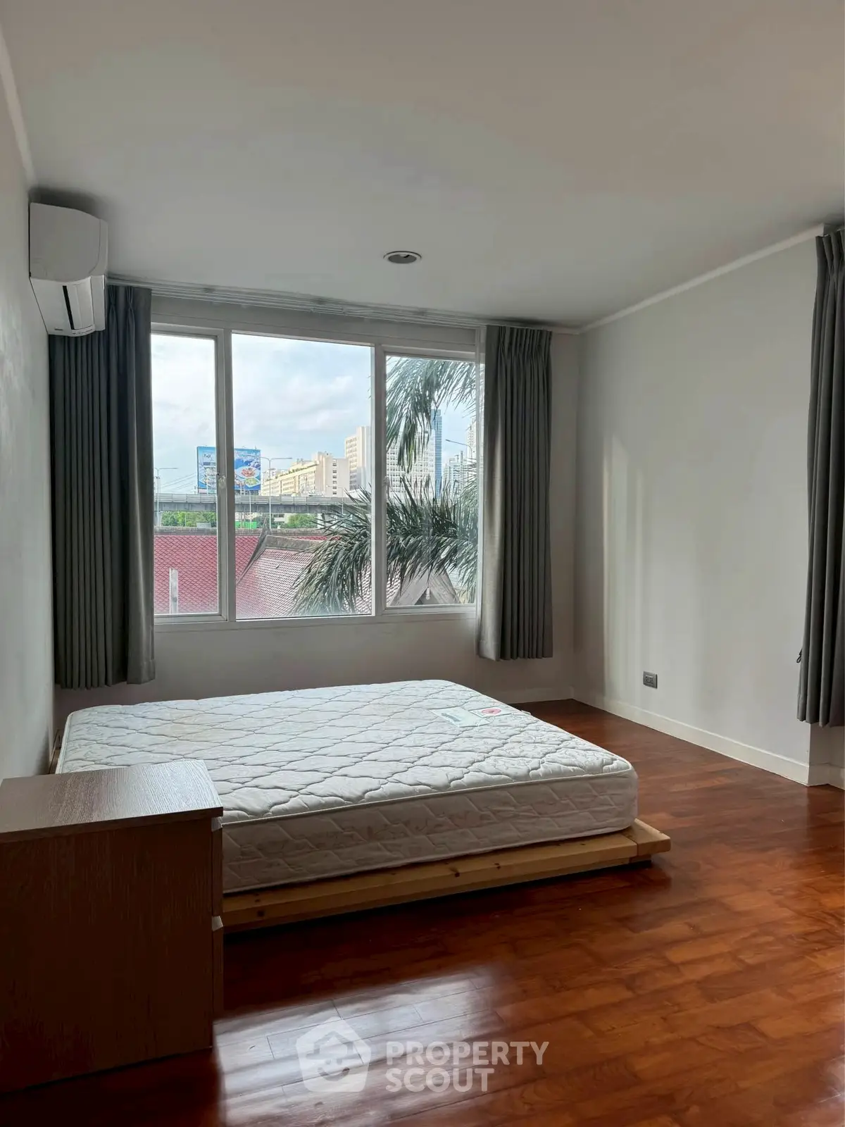 Bright bedroom with large window and wooden flooring, offering a city view and natural light.