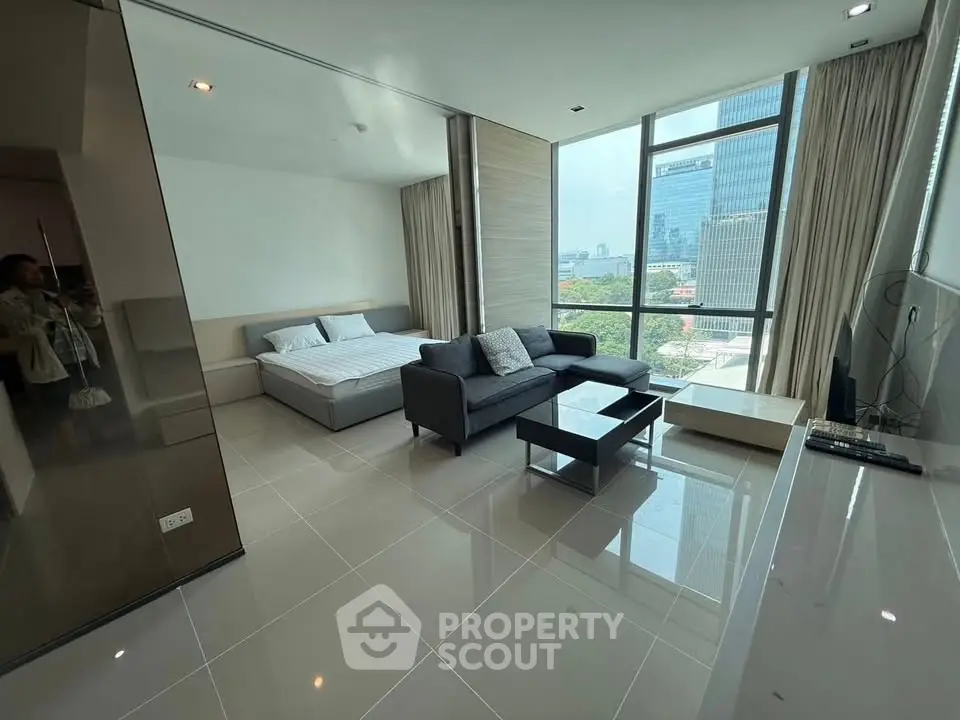 Modern studio apartment with large windows and city view, featuring a cozy bed and stylish sofa.