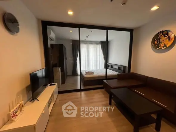 Modern living room with glass partition to bedroom, featuring sleek furniture and wooden flooring.